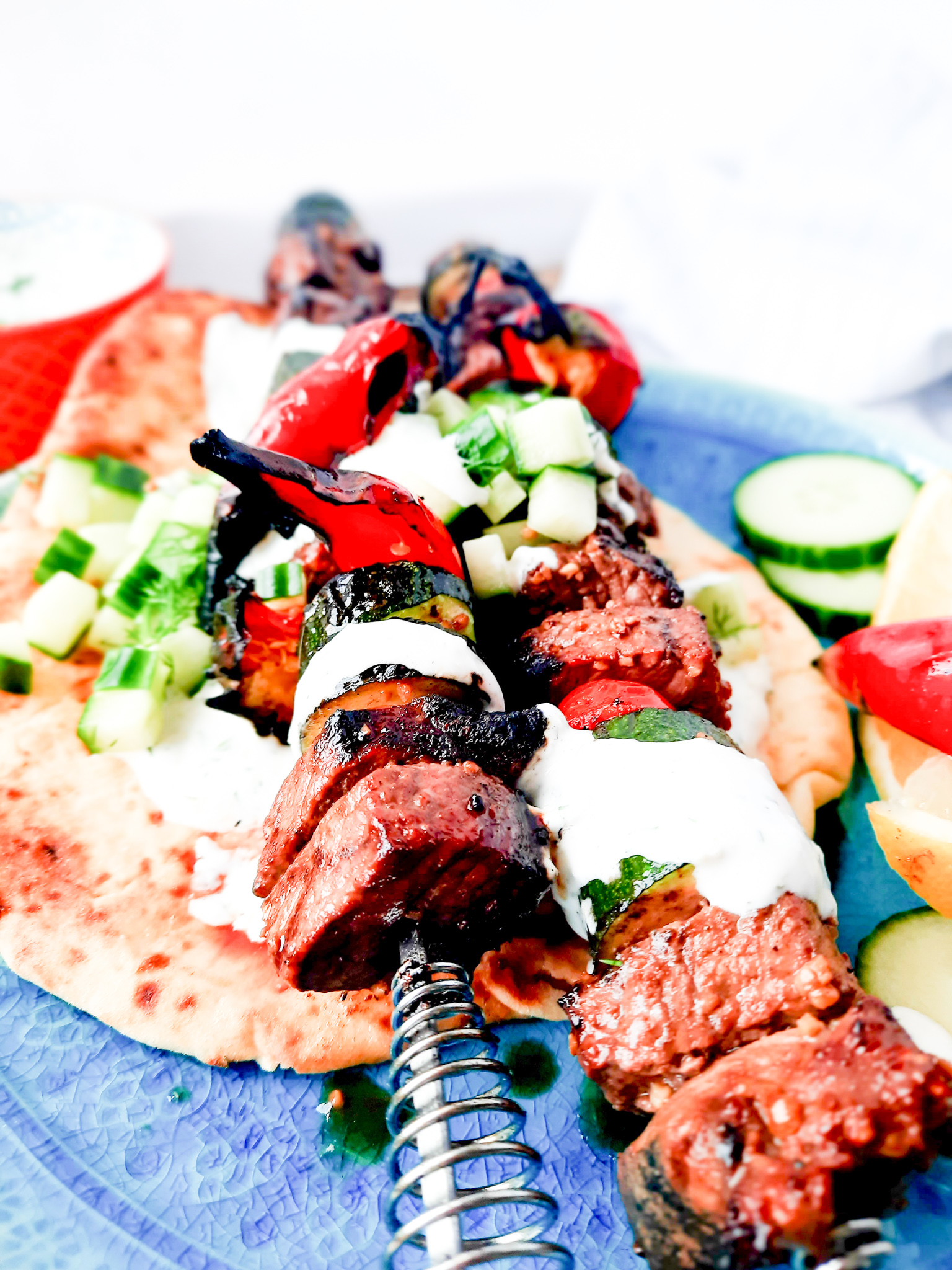 Tender, marinated Greek Inspired Steak Kabobs with Tzatziki, zucchini, mushroom, chili peppers and cucumber dill relish top pita bread.