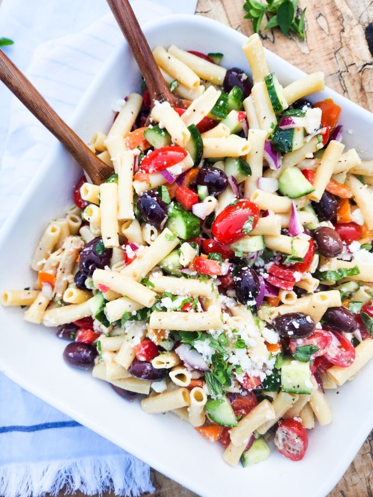 A colorful pasta salad featuring penne pasta mixed with cherry tomatoes, cucumbers, bell peppers, red onion, black olives, and feta cheese, served in a white dish with wooden serving utensils.