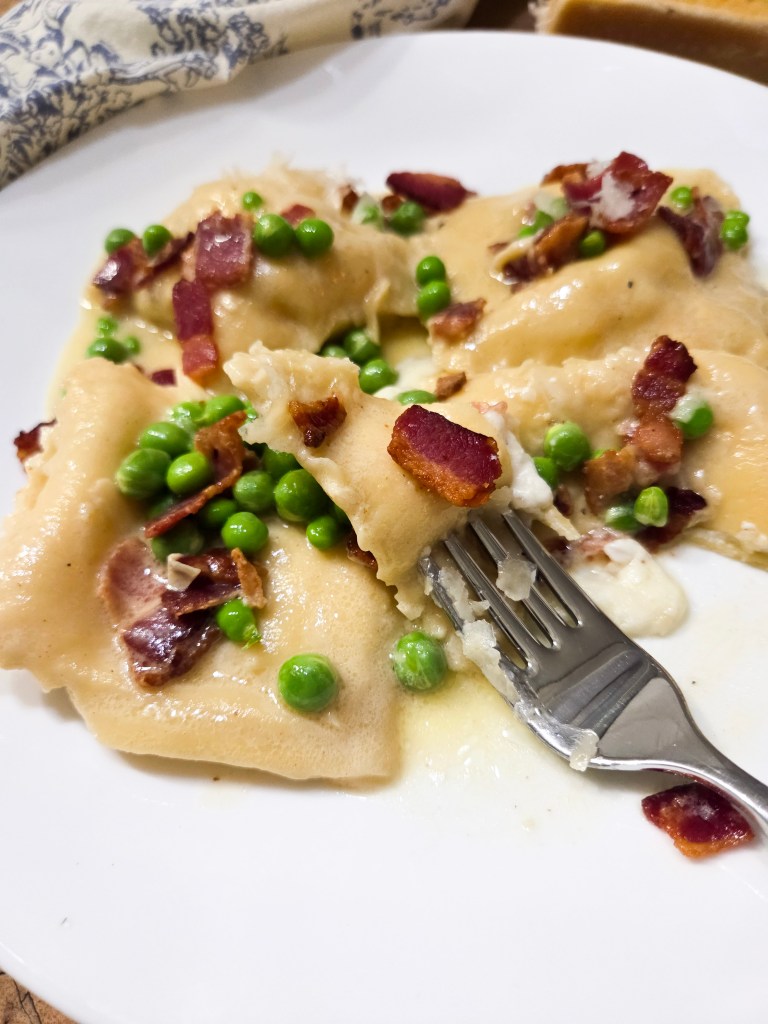 A plate of ravioli topped with crispy bacon and green peas, served with a fork partially lifting a piece.