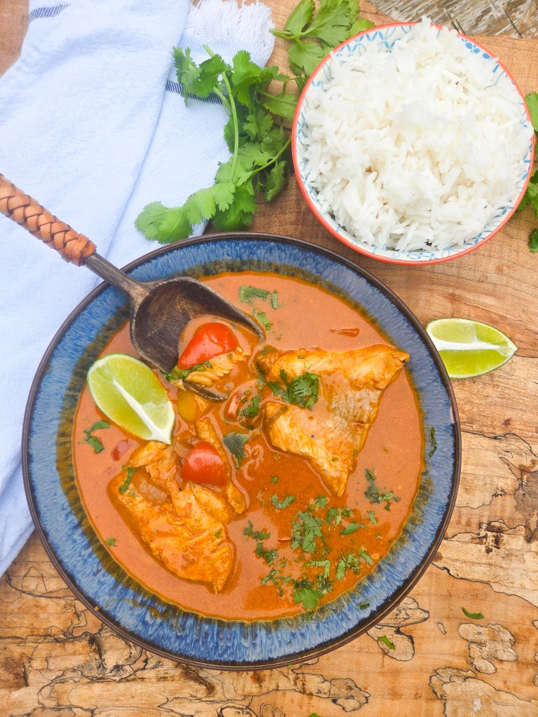 A bowl of fish curry garnished with cilantro and lime, accompanied by a side of white rice in a decorative bowl.