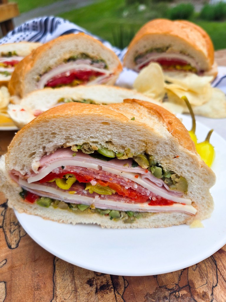 A close-up of a sliced sandwich on a white plate, showcasing layers of ham, salami, sliced pickles, and red peppers between two pieces of bread, with potato chips and pickled peppers on the side.