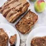A loaf of apple cider bread with slices on plates and whole apples near the edge