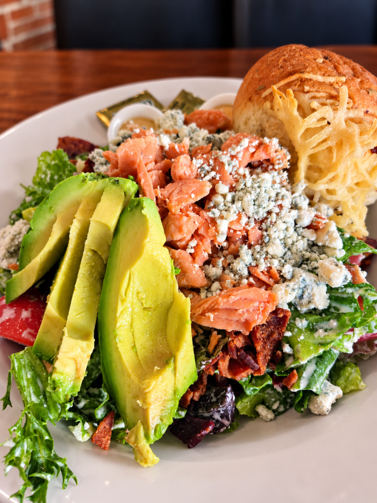 A colorful salad topped with slices of avocado, smoked salmon, crumbled blue cheese, and bacon, served with a soft roll on the side.