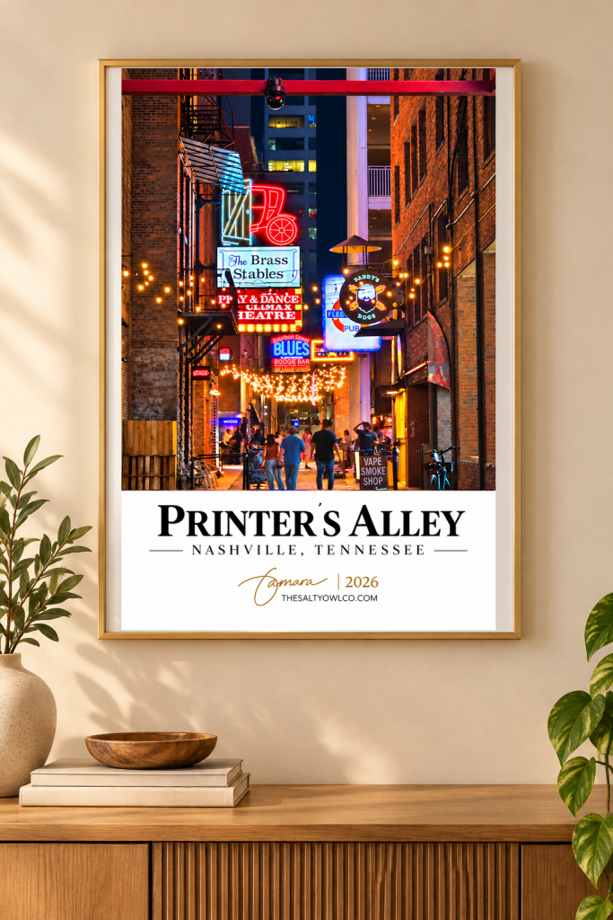 A framed poster of Printer's Alley in Nashville, Tennessee, featuring vibrant neon signs and string lights illuminating a bustling alley with people socializing.