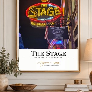 The Stage Nashville travel poster in a slim gold frame on a cream-paneled wall above a rustic wooden console, accented with greenery, warm lighting, and woven furnishings.