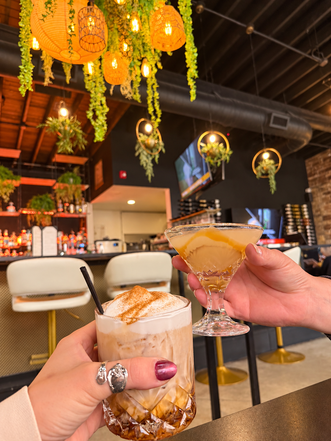 Two fall cocktails held in front of the warmly lit bar at 1933 Bar+Kitchen.