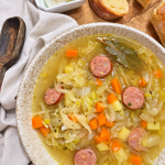 Overhead view of Polish sauerkraut soup with sausage, potatoes, and carrots, served with bread and sour cream.